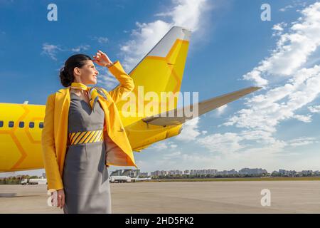 Allegro assistente di volo in uniforme della compagnia aerea che copre gli occhi dal sole con la mano e sorride mentre si alza contro il cielo nuvoloso Foto Stock