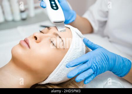 Cura professionale della pelle. Cosmetologist in guanti blu fa ecografia pulizia del viso di una donna con scrubber Foto Stock