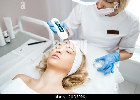 Cosmetologist o dermatologist che fanno la pulizia facciale ad ultrasuoni per la donna in salone di bellezza. Procedura per la cura della pelle Foto Stock