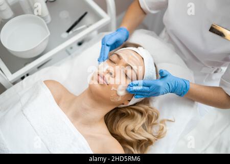 Medico estetista in guanti blu sta applicando schiuma o crema per la pulizia del viso sulla pelle del viso della donna massaggiante in clinica di cosmetologia. Femmina beauty concep Foto Stock