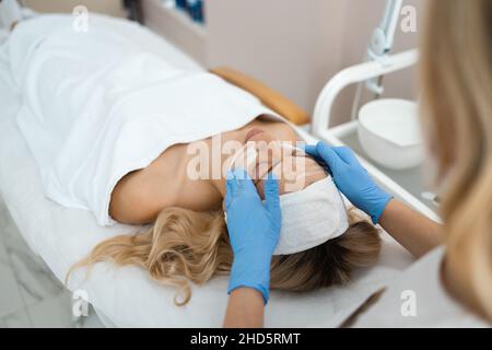 Medico cosmetologo applica una maschera di schiuma detergente e idratante al volto di una bella donna. Giovane bionda alla procedura di spa Foto Stock