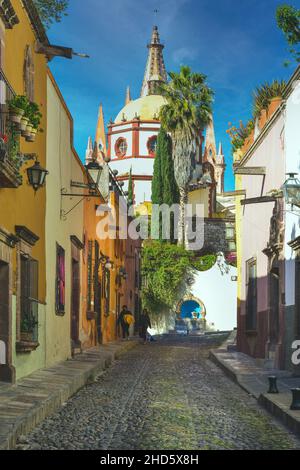 La stretta Cobblestone Aldama Street offre la vista della cupola della Chiesa di Parroquia San Miguel de Allende Arcangel accanto a case colorate in SMA, Messico Foto Stock