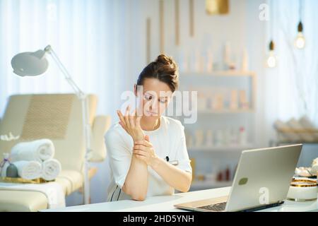 lavoratore donna stanca con computer portatile che ha dolore al polso in salone di bellezza moderno. Foto Stock