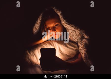 Giovane donna sdraiata a letto e con smartphone durante la notte prima di dormire. Dipendenza dai social media e insonnia Foto Stock