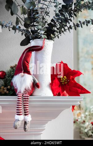 Una classica vacanza norweigan gnome decorazione di Natale seduta su un bianco manto moderno con un vaso bianco con eucalipto e una poinsettia fiore Foto Stock