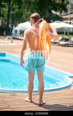 Un uomo in pantaloncini blu in piedi vicino alla piscina con un tubo in mano Foto Stock