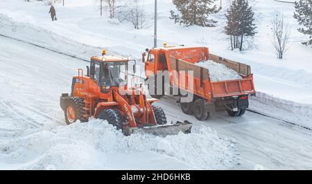 Dicembre 23, 2021. Regione di Kemerovo, Russia. Il trattore arancione grande pulisce la neve dalla strada e la carica nel carrello. Pulizia e pulizia della strada Foto Stock