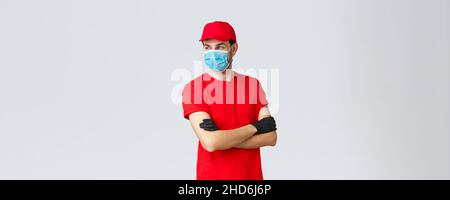 Covid-19, auto-quarantena, shopping online e concetto di spedizione. Corriere sicuro in uniforme rossa, guanti protettivi e maschera facciale, in piedi incrociati Foto Stock