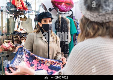 Dipendente di un negozio di abbigliamento di seconda mano che mostra abiti a un cliente, lavorando con misure di sicurezza nella pandemia di coronavirus Foto Stock