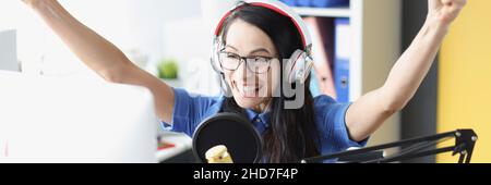 Una donna sorridente gioiosa solleva con entusiasmo le mani in cuffia e nel microfono e guarda il monitor del computer Foto Stock