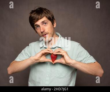 Giovane uomo bianco sottile, alto e bello, con capelli marroni che fanno forma al cuore con le mani che tengono la fragola in una camicia blu chiaro su sfondo grigio Foto Stock