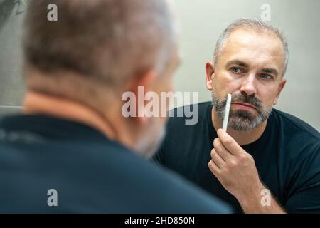 Un bell'uomo di mezza età che gli spazzolava la barba la mattina in bagno guardando nello specchio Foto Stock