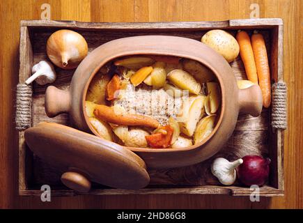 Vista dall'alto della pentola di terracotta con arrosti di maiale e verdure cotte lentamente all'interno su vassoio di legno e tavolo di legno, circondato da biologico crudo. Foto Stock