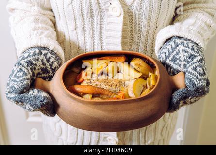 Donna che tiene in mano la pentola di cottura in terracotta con arrosti di maiale e verdure cotte lentamente all'interno. Indossare abiti a maglia, concetto di comfort invernale. Foto Stock