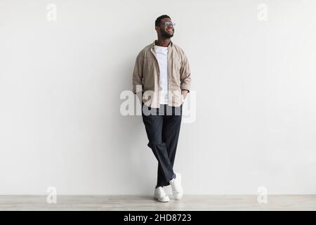 Tutta la lunghezza del giovane afro-americano bello uomo che indossa abiti casual, guardando da parte, posando contro il muro bianco Foto Stock