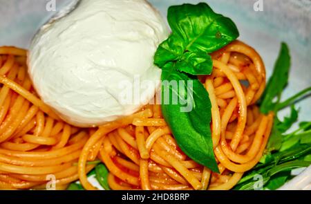 Basilico a foglia verde accanto alla burrata, palla di mozzarella con un ripieno di listini di formaggio, crema di formaggio e panna, su spaghetti cotti con salsa di pomodoro. Foto Stock