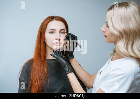 Il padrone estetician fa il markup del sopracciglia che tatuing, le donne prima di applicare il trucco permanente, il trucco permanente dell'occhio e la procedura di correzione del sopracciglio. La ragazza felice del cliente è molto soddisfatta Foto Stock