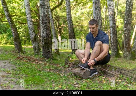 Uomo che lega le sue scarpe nel parco all'aperto, intorno alla foresta, querce verde erba verde giovane atleta resistente atleta. Foresta sportiva, percorso di allenamento fi Foto Stock