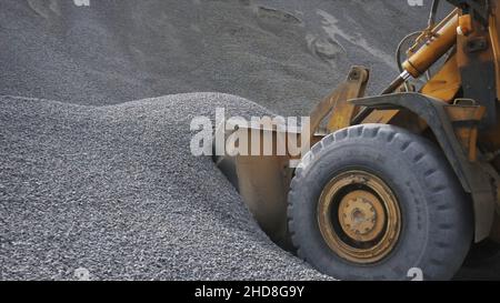 Il trattore raccoglie una paletta con ghiaia. Vista laterale di un escavatore che carica la benna con pietre o macerie schiacciate nel cantiere. Foto Stock