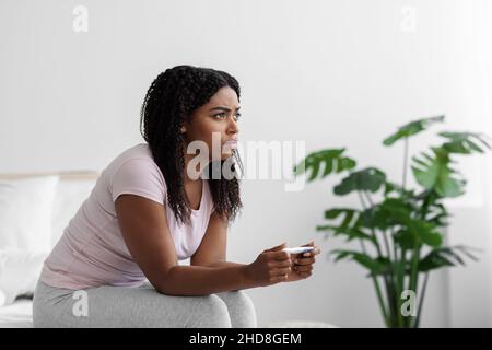 Frustrata triste giovane donna nera seduta a letto e guardando il test di gravidanza negativo hanno problemi di salute femminile Foto Stock