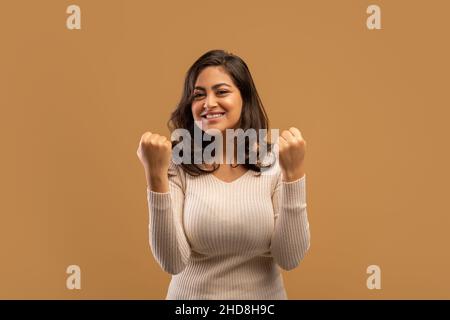 Concetto di vincita. Una donna araba gioiosa che festeggia la vittoria, scuotendo i pugni arroccati sullo sfondo beige dello studio Foto Stock