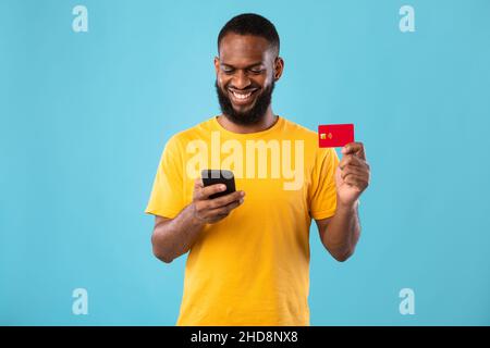 Positivo ragazzo nero giovane con carta di credito e smartphone, shopping online su sfondo blu studio Foto Stock