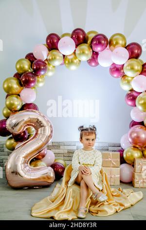Bambina alla moda in un abbigliamento bianco seduto su sedia, sorridente e festeggiante il suo secondo compleanno. Bella foto compleanno ragazza bambino Foto Stock
