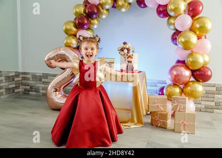 Divertente bambina di due anni con vestiti rossi che si divertono di fronte alla sua torta di compleanno. Bella foto compleanno ragazza bambino Foto Stock