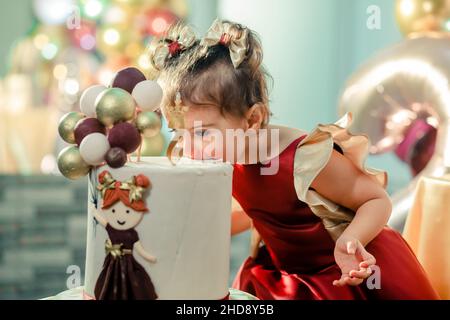 Bella, dolce bambina in un abito rosso di lusso morde la sua seconda torta di compleanno. Bella foto compleanno ragazza bambino Foto Stock