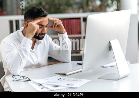 Giovane indiano adulto sconvolto, proprietario dell'azienda, direttore o lavoratore di ufficio con la barba, siede alla sua scrivania in ufficio moderno con gli occhi chiusi, gli sfrega la testa, si sente stressato e stanco dal lavoro straordinario, ha bisogno di riposo Foto Stock