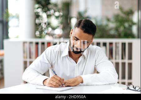 Concentrato di uomo d'affari indiano serio e sicuro, responsabile o freelance, indossando una camicia bianca, seduto al tavolo in un ufficio moderno e prendendo appunti, ascoltando il webinar online, brainstorm Foto Stock