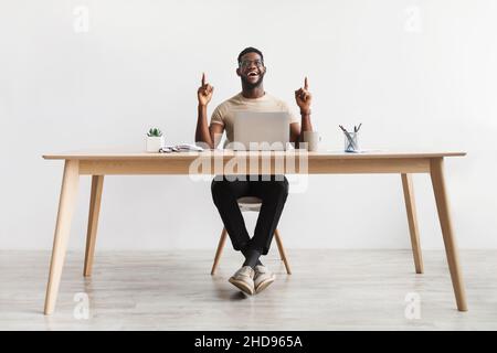 Millennial black Office lavoratore avere idea creativa, gesturing eureka sul posto di lavoro, seduto a tavola contro muro bianco Foto Stock