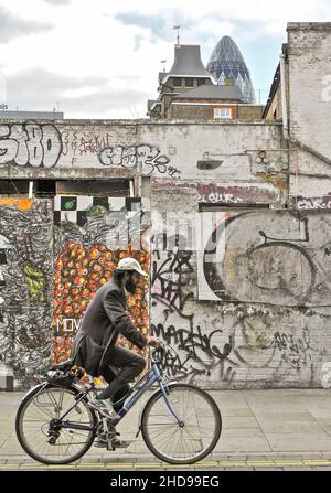 Un uomo asiatico in bicicletta lungo Brick Lane, nell'East End di Londra, con il grattacielo Gherkin sullo sfondo. Foto Stock