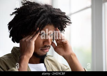 Giovane ragazzo nero sconvolto che soffre di mal di testa mentre si sta in piedi vicino alla Grande finestra Foto Stock