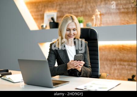 Gioioso successo attraente influente maturo caucasica donna d'affari o top manager utilizzando il cellulare mentre si siede alla scrivania in ufficio, inviare messaggi di testo a clienti, amici o colleghi, ha ottenuto buone notizie, messaggi Foto Stock