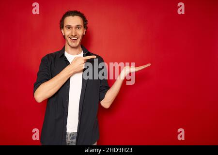 Giovane, bello, uomo eccitato in abiti casual, maglietta blu, T-shirt bianca, punti indice dito verso mano libera spazio vuoto per pubblicità testo oggetto isolato rosso sfondo copia spazio. Vendita, promo. Foto di alta qualità Foto Stock