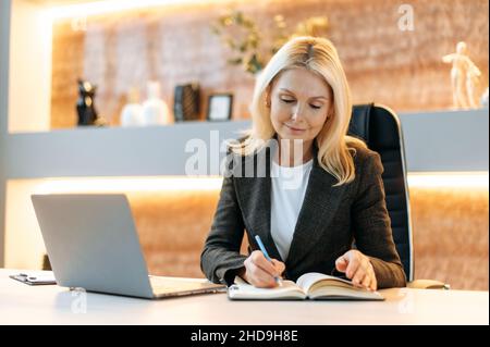 Successo intelligente attraente influente maturo caucasico bionda donna d'affari, ceo o agente immobiliare seduta in ufficio in tuta, utilizzando il laptop, prendere appunti, ponderare la strategia di business, sorridente Foto Stock