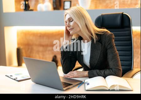 Stravagata donna d'affari matura o top manager, in tuta, stanco di lavorare online su un laptop, di sperimentare stress e mal di testa, di prendersi pausa dal lavoro, massaggiare il suo tempio, ha bisogno di riposo, occhi chiusi Foto Stock