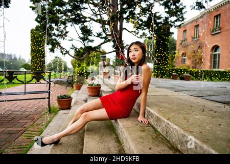 Giovane donna dell'Asia orientale in Red Party / vestito di festa che si rilassa su gradini giocando con le luci Foto Stock