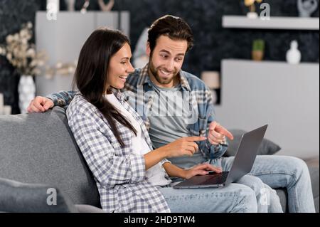 Coppia caucasica positiva che usa il laptop mentre si riposa sul divano in salotto a casa, passare il tempo insieme, navigare sui social media, internet, guardare video, messaggi con gli amici, sorridenti Foto Stock