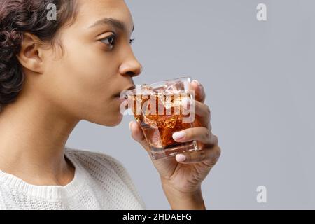 Primo piano ritratto di giovane donna afroamericana che beve cola con ghiaccio su sfondo grigio Foto Stock