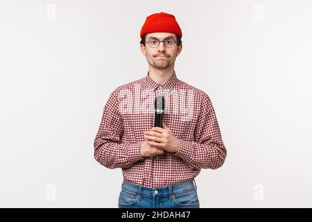 Ritratto ravvicinato di timido e geeky giovane ragazzo bearded in occhiali e beanie rosso, guardando lo schermo tv e tenendo il micrphone, prepararsi a cantare Foto Stock