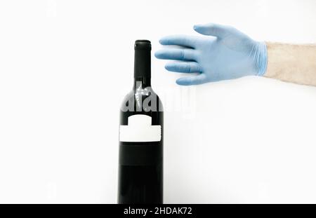 Mano con guanti blu che raggiungono la bottiglia di vino rosso su sfondo bianco. Norme di sicurezza e industria delle bevande durante il concetto di pandemia da virus corona. Foto Stock