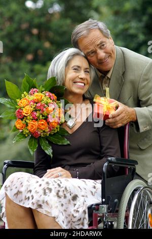 Ein aäteres Paar im Park - die Frau sitzt im Rollstuhl, der Mann umarmt sie liebevoll und ueberreicht ein Geschenk und Blumen, Model-Release| Vecchio colpo di Stato Foto Stock