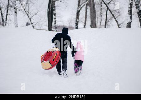 Foto lontana di femmina capretto e padre con slitta che salirà su collina per scivolare giù nella neve nella foresta. Sfondo sorprendente pieno di bianco e neve Foto Stock