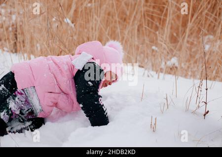 Primo piano foto di femmina russo ragazzo strisciando sulla neve e guardando la neve indossare abiti rosa inverno nella foresta. Sfondo sorprendente pieno di bianco Foto Stock