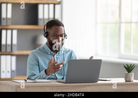 Allegro attraente maschio nero millenario con barba in occhiali, cuffie gesti con le mani guardare il pc Foto Stock