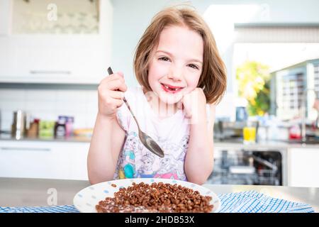 Una giovane ragazza con due denti incisivi anteriori decidui mancanti e due denti incisivi secondari anteriori che iniziano a crescere mangia una ciotola di cereali da colazione cocopos Foto Stock