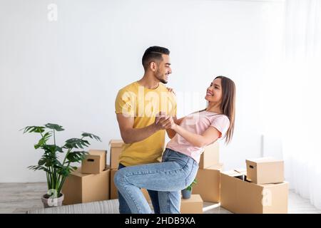 Felice millennial diverse coppia ballare nella loro nuova casa tra scatole di cartone il giorno di trasferimento, spazio copia Foto Stock