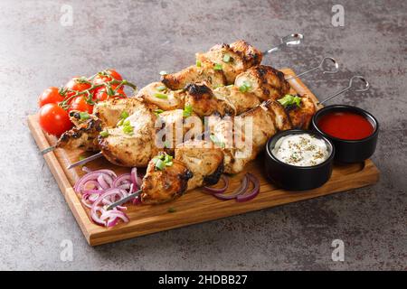 Gustosi e succulenti spiedini di carne alla griglia con verdure fresche e due salse da vicino su un vassoio di legno su sfondo grigio di cemento. Orizzontale Foto Stock